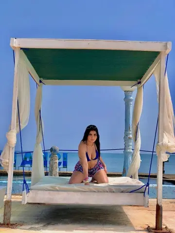 Young woman in a blue bikini relaxing on a white cabana bed by the ocean under a clear blue sky.