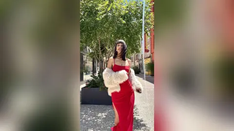 Smiling woman in a red evening gown and white faux fur coat walking on a sunny city street.