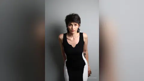 IveteDurand poses in a tight black and white tank top dress against a plain grey background, showing off her short dark hair and arm tattoo.