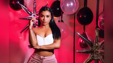Glamorous woman in a white crop top posing amidst pink and black balloons in a festive party setting.
