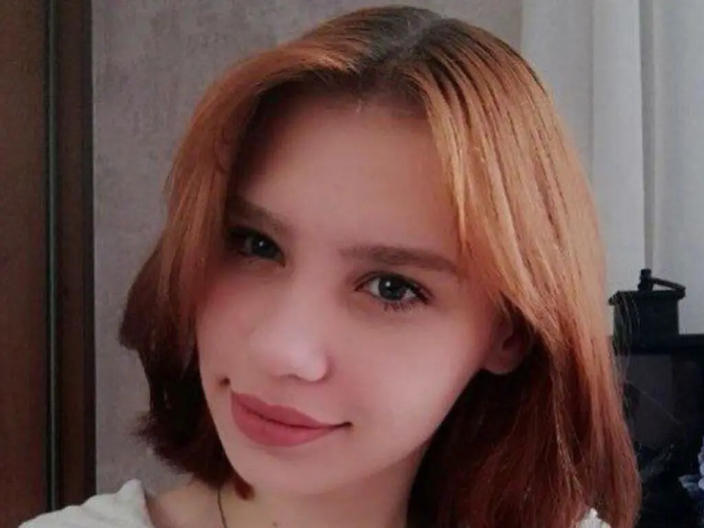Smiling young woman with reddish-brown hair in a casual white top indoors.