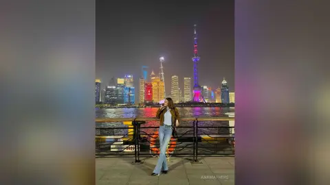 Young woman in a brown jacket and jeans standing by a river overlooking the illuminated Shanghai skyline at night.