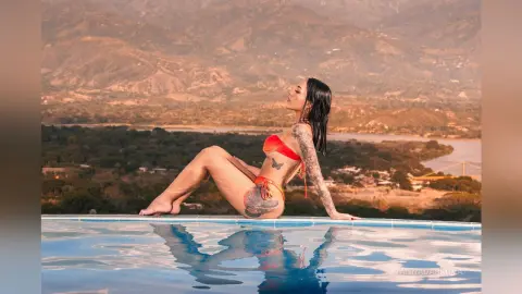 Woman in orange bikini relaxing by an infinity pool overlooking a scenic mountain valley at sunset.
