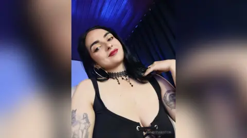 Woman with dark hair and tattoos wearing a black tank top and lace choker in moody blue lighting.