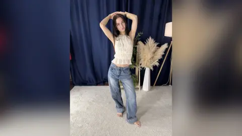 Young woman in a white ruffled halter top and baggy blue jeans posing against a navy blue backdrop.