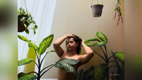 A woman in a green leaf top poses amidst lush tropical plants in a bright, sunlit indoor garden setting.