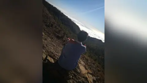Man in a blue t-shirt standing on a rocky mountain slope overlooking a vast valley under a bright sky.