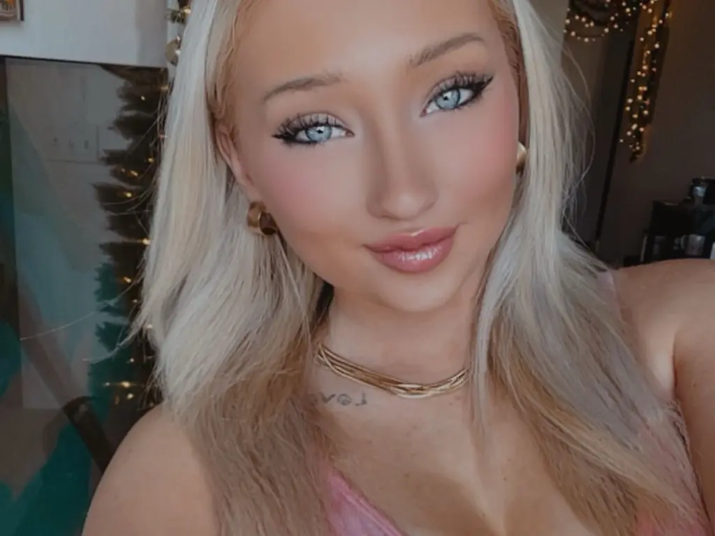 Avaquinnxo poses with a soft smile, showing her long blonde hair and light blue eyes while wearing a pink top.