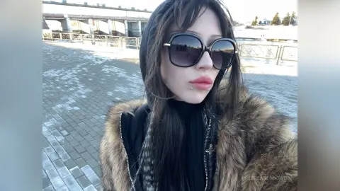 Young woman in a black hoodie and brown faux fur coat wearing sunglasses on a sunny urban walkway.