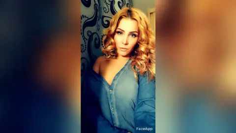 Blonde woman in a blue denim shirt posing against a patterned blue wallpaper in a moody indoor setting.