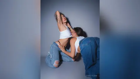 Two women in white tops and denim jeans posing playfully against a grey studio background.