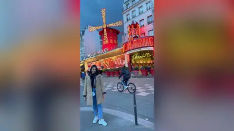 Woman in a beige trench coat standing on a street in front of the illuminated Moulin Rouge in Paris at night.