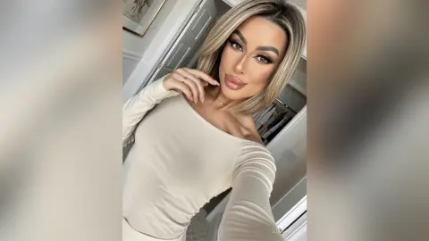 SinnaRay poses in a white long-sleeve top with her blonde hair styled down, looking into the camera.