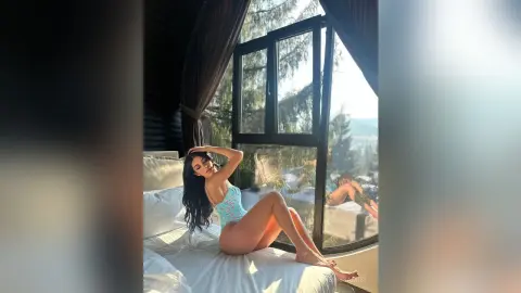 NerissaMist sits on a white bed in a light blue bodysuit, posing by a large window with a view of trees.
