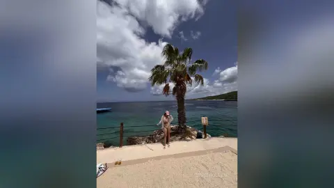 RossiNoleen stands naked on a sandy beach near the ocean, framed by a single palm tree under a cloudy sky.