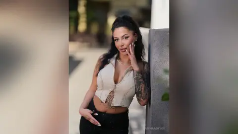 Stylish woman in a white cropped vest and black jeans leaning against a wall in soft outdoor lighting.