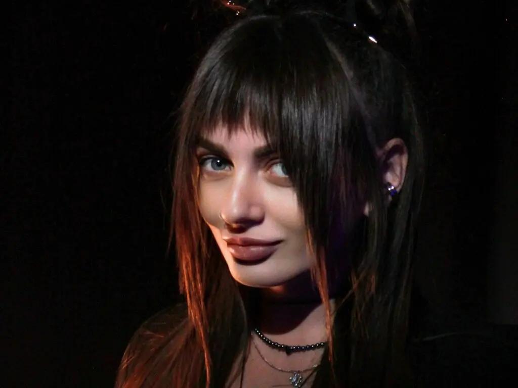 JaneMabbie looks into the camera with long dark hair and bangs in a dimly lit setting.