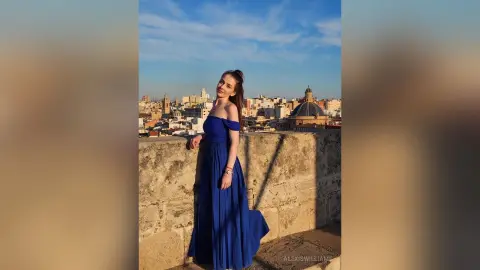 Elegant woman in a long blue evening gown posing on a stone balcony overlooking a historic European city skyline.
