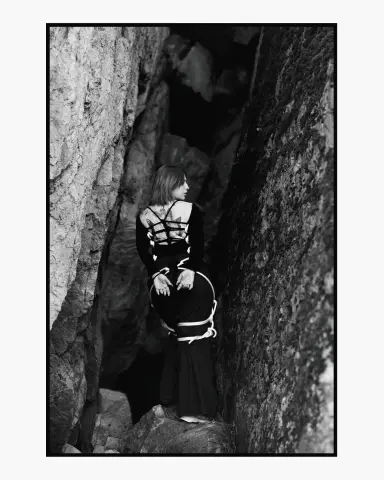 AlisaMoss stands in a dark rocky crevice wearing a black outfit with white straps around her torso and legs.