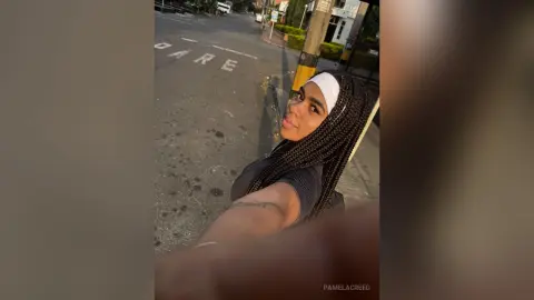 PamelaCreed poses outdoors with her long dark braids and white headband, wearing a black top while looking at the camera.