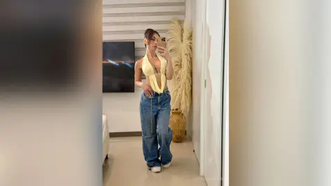 Young woman in a yellow crop top and baggy jeans taking a mirror selfie in a modern minimalist room.