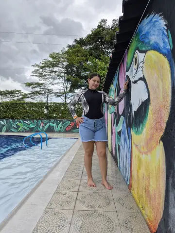 Smiling woman in a black bodysuit and denim shorts standing by a colorful parrot mural next to a swimming pool.