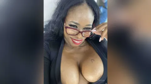 KattyLeya — KattyLeya smiles wearing red glasses and a low-cut black top that shows her cleavage.