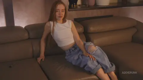 AddieRunk sits on a brown leather couch wearing a white tank top and ripped blue denim jeans, letting her long light-brown hair hang down.