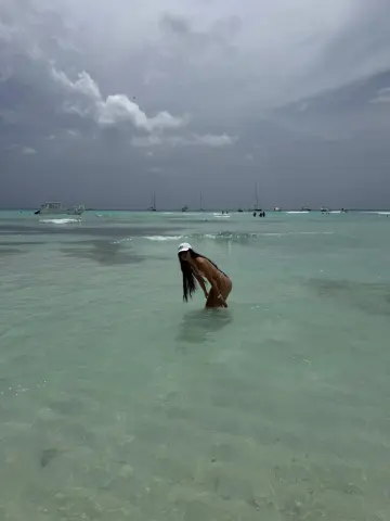 Young woman in a white bikini wading in turquoise ocean water under a cloudy sky at a tropical beach.