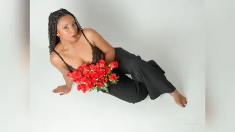 Elegant woman in a black lace camisole and trousers holding a bouquet of red roses against a white background.