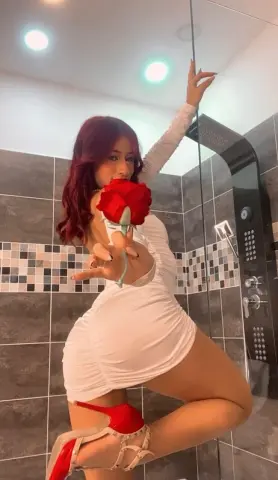 Young woman in a white mini dress holding a red rose in a modern tiled bathroom.