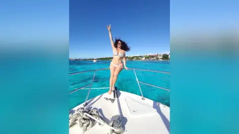 Cheerful woman in a white bikini posing on a boat deck amidst turquoise tropical ocean waters under a clear blue sky.