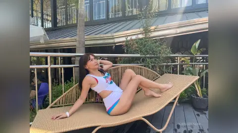 Young woman in a white crop top relaxing on a wicker lounge chair on a sunny outdoor wooden deck.