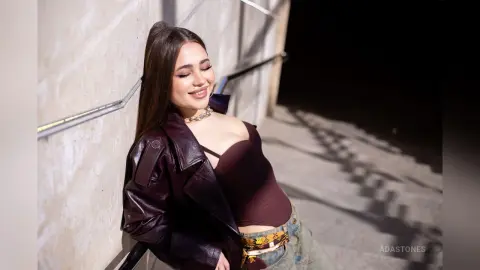 Smiling young woman in a dark leather jacket and crop top leaning against a wall at night.