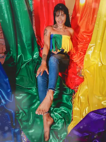 EmmaEbonyX19 smiles while sitting against rainbow-colored satin drapes, wearing a colorful fringe tube top and tight blue jeans.