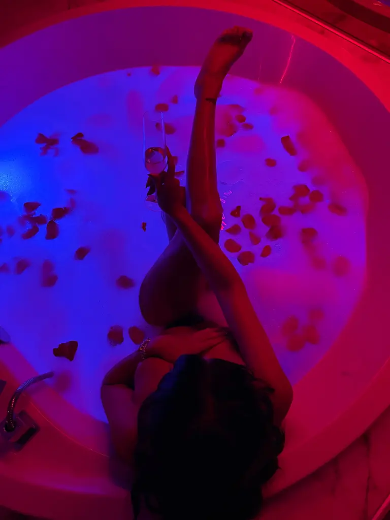 KimRiver — Relaxed woman in a rose petal bathtub with blue and pink neon lighting for a moody spa aesthetic.