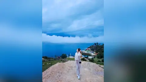 LaurentBelluci stands on a dirt path overlooking a blue sea and green hills, wearing a white top and light grey pants.