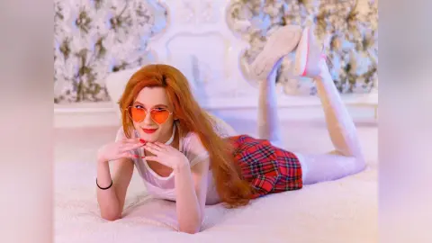 Redheaded woman in a white t-shirt and red plaid skirt relaxing on a bed with a playful mood.