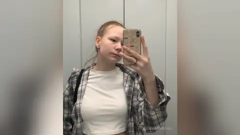 Young woman in a white t-shirt and grey plaid shirt taking a casual mirror selfie in a bright room.