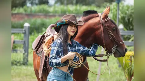 JessieLiu wears a brown cowboy hat and blue plaid shirt while posing next to a brown horse in a grassy field.