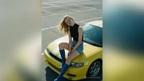 Stylish blonde woman in a black sleeveless top and denim shorts sitting on a yellow car in a sunny parking lot.