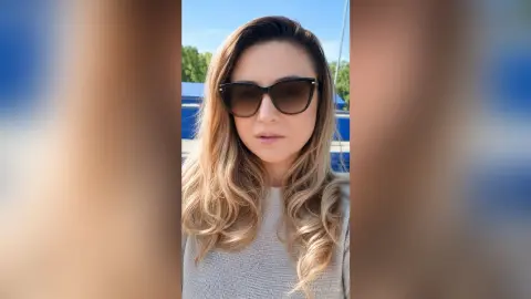 MaliaQuinn wearing large dark sunglasses and a light grey knit sweater, posing outdoors with long blonde hair.