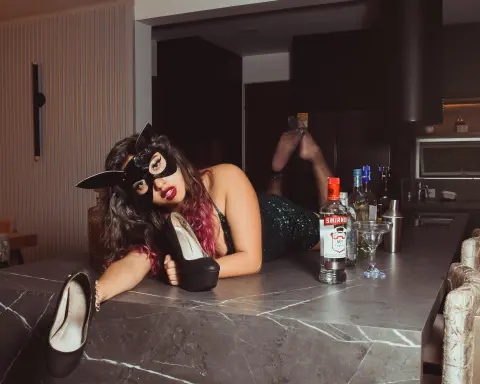Woman in a black bunny mask and sequin outfit lounging on a marble kitchen counter with liquor bottles.