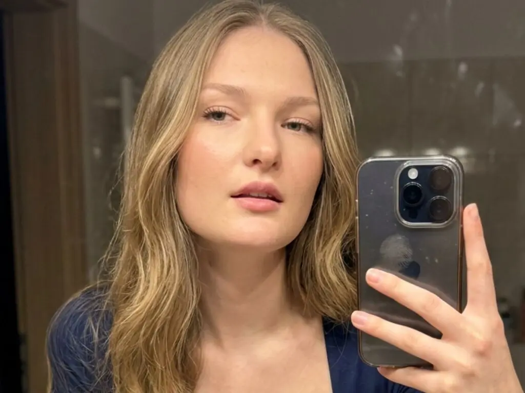Blonde woman in a blue top taking a casual selfie in a dimly lit room.