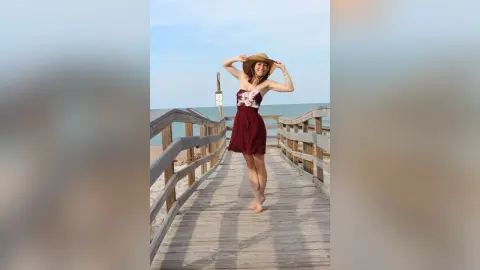Cheerful woman in a burgundy floral sundress and straw hat walking on a sunny wooden beach pier.
