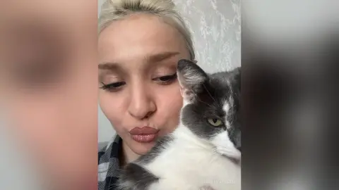 BarbaraGreen pouts her lips while holding a grey and white cat close to her face against a light grey wall.