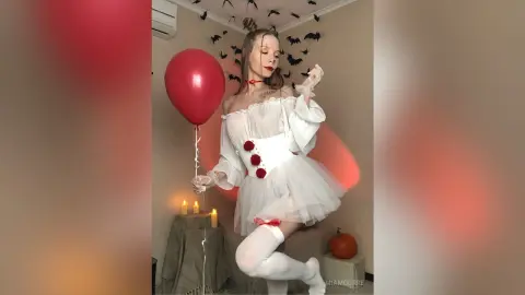Woman in white gothic Lolita costume holding a red balloon in a spooky Halloween room with bats and pumpkins.