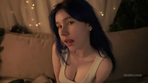 Young woman with blue hair in a white tank top relaxing in a cozy, dimly lit bedroom setting.