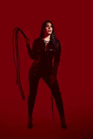 Confident woman in a black leather jumpsuit holding a whip against a bold red background.