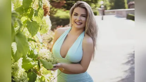 Sofiya smiles while wearing a light blue halter-neck top and matching skirt, standing next to white flowering bushes outdoors.